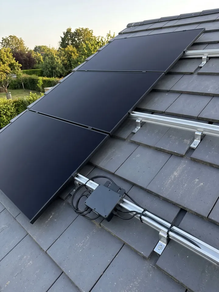 Des panneaux photovoltaïques noirs sur des tuiles grises, vus de près, les rails et un micro-onduleur sous un panneau. Illustration.
