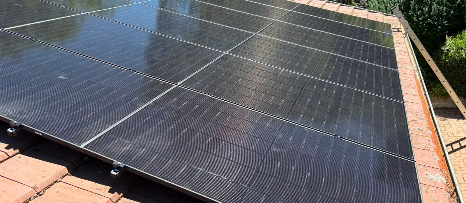 Trois rangées de panneaux photovoltaïques noirs sur une toiture en tuiles, jardin et haies en contrebas